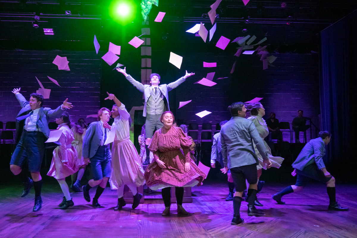 2022 Spring Awakening Promotional Photo 1 Boy standing on a desk tossing papers into the air as others dance around him