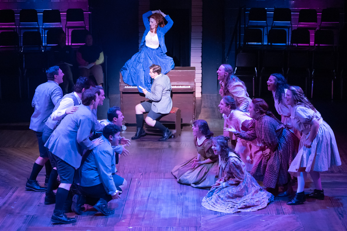 2022 Spring Awakening Promotional Photo Woman on top of a piano as a group of boys and a group of girls in the forefront sing